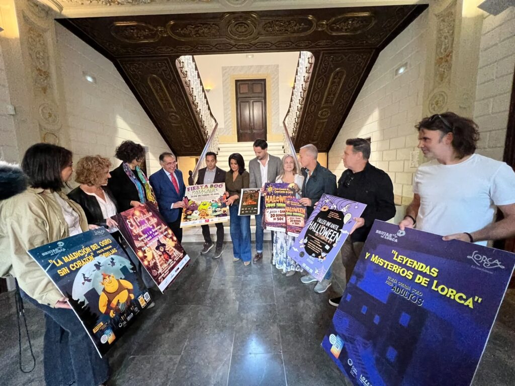 Participantes en la presentación de la programación de Halloween en Lorca muestran los carteles de las actividades en el vestíbulo de un edificio histórico.